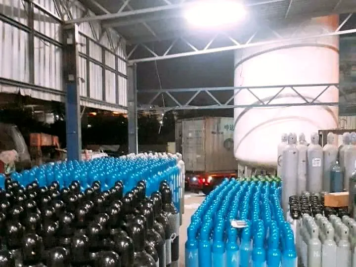 Many blue and black gas cylinders lined up in a warehouse with a large white storage tank behind